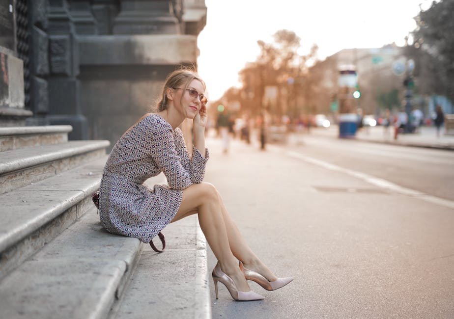 A fashionable woman in a patterned dress sits on steps during sunset, city street life around her.